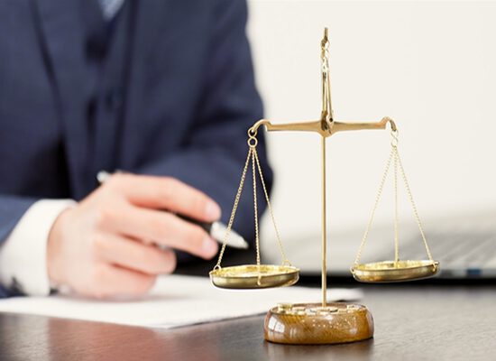法律の象徴である天秤と書類に向き合う弁護士の手元。法的判断の慎重さを示す。
